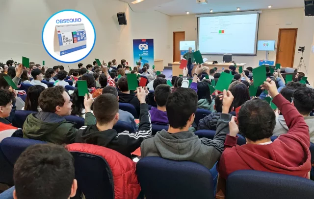 Estudiantes en el salón de actos levantando tarjetas verdes y rojas, en una actividad gamificada. Se muestra en un círculo un calendario como obsequio para los asistentes