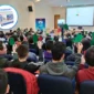 Estudiantes en el salón de actos levantando tarjetas verdes y rojas, en una actividad gamificada. Se muestra en un círculo un calendario como obsequio para los asistentes